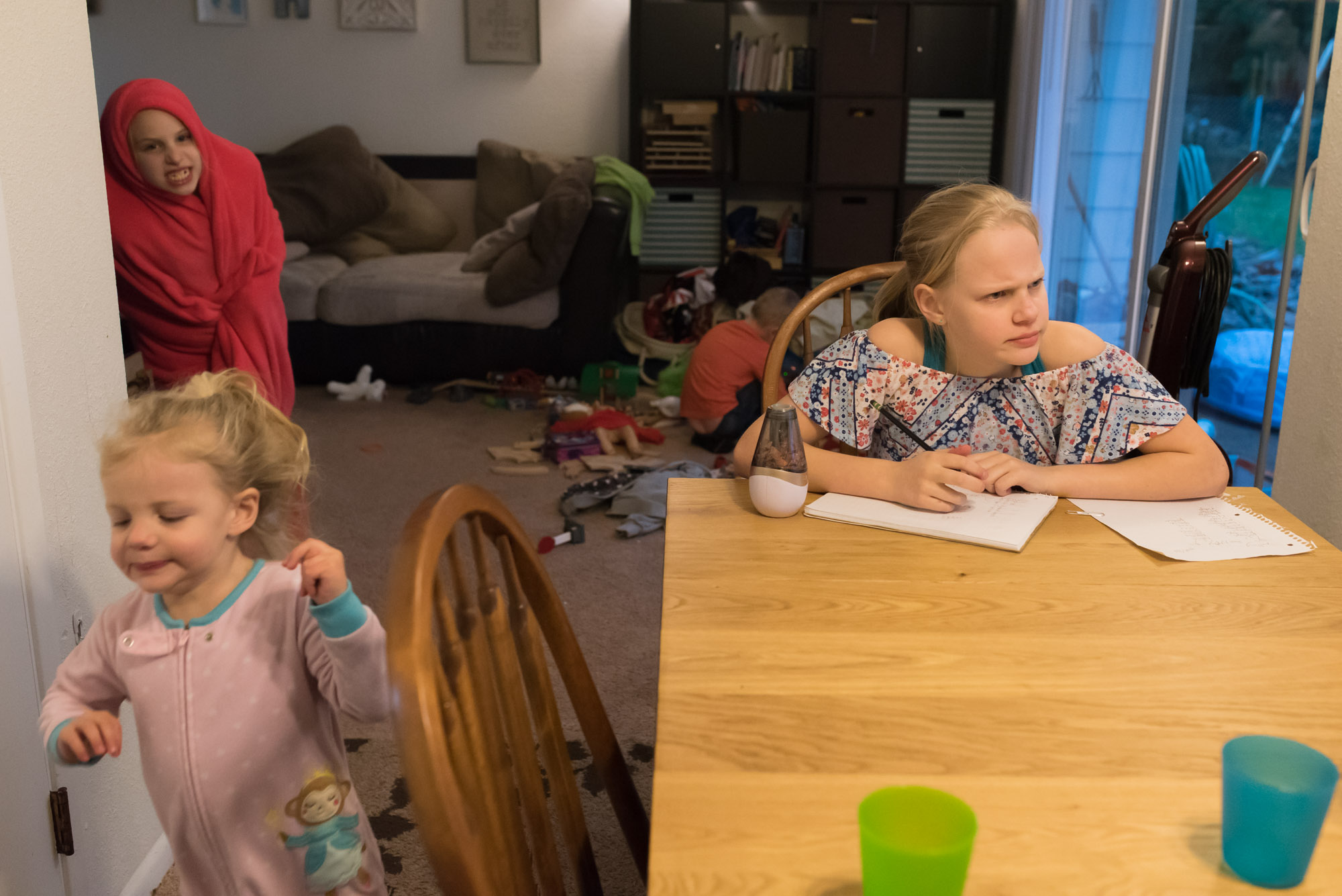 girl does homework while younger siblings play wildly - documentary photography military families