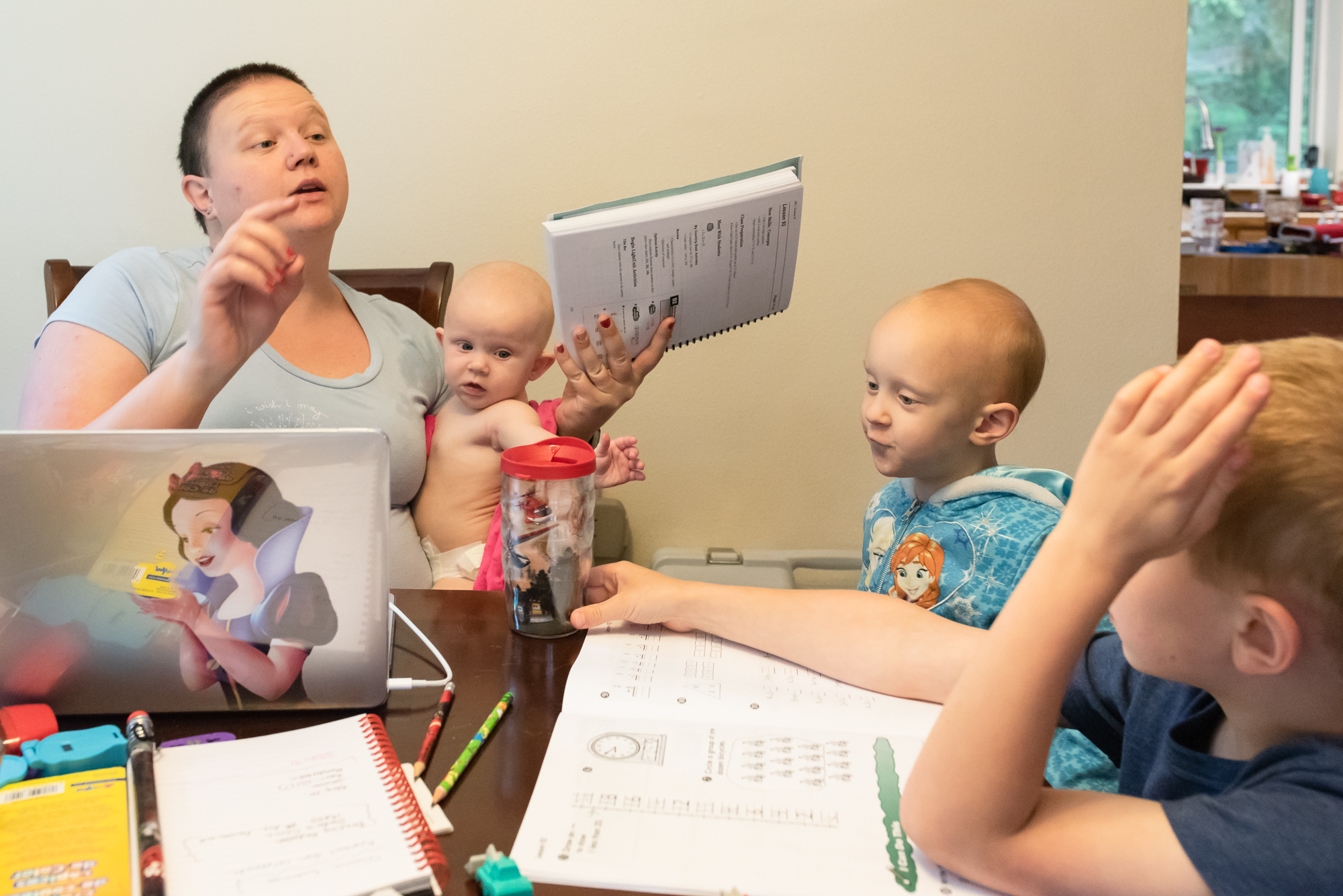 mother does school work with son with baby in lap and sister stands nearby